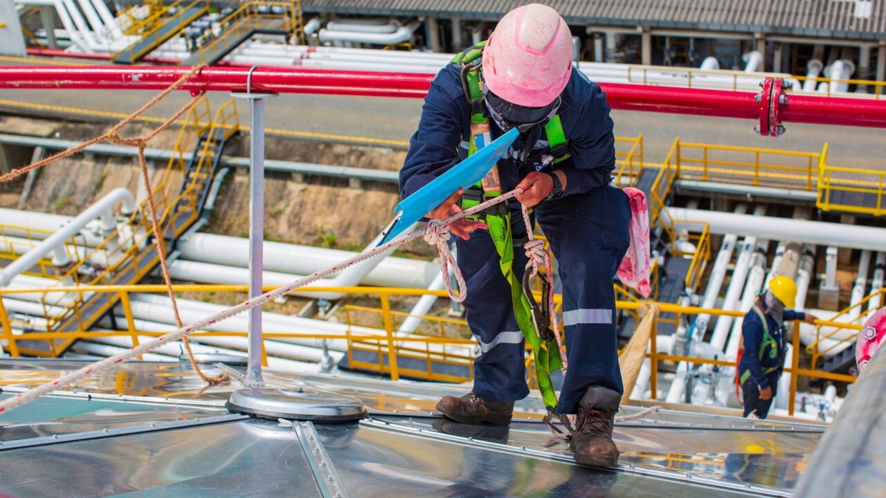 Worker in safety gear adjusts harness on a sloped metal surface with industrial pipes and structures in the background.