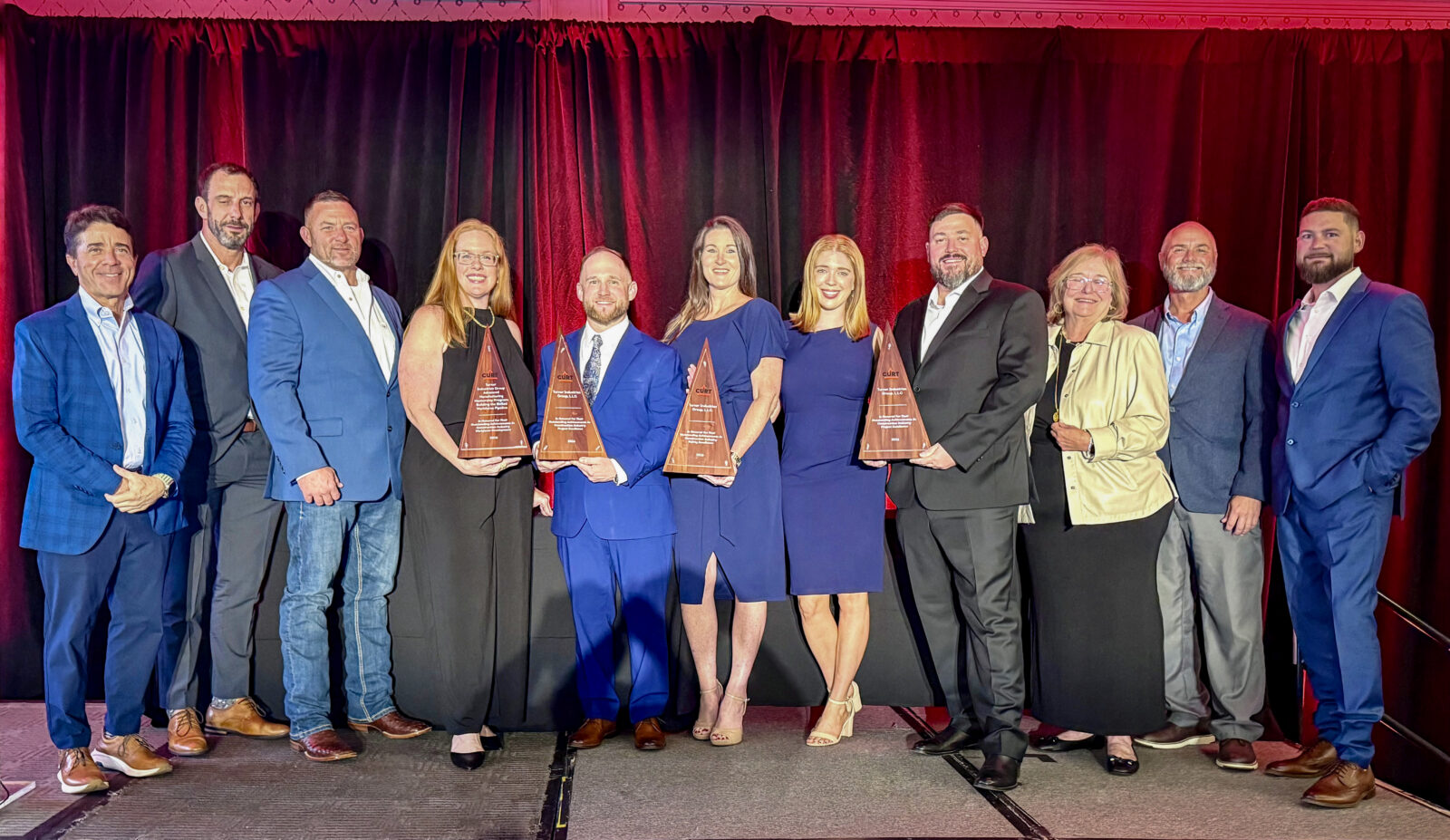 A group of ten people stand in a line on a stage, with four people in the center holding triangular glass awards.